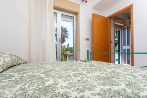 A bedroom with a bed, a door leading to a balcony, and a view of palm trees outside.