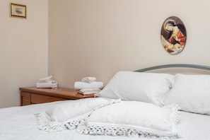 Un letto rifatto con lenzuola bianche e un cuscino decorativo, un comodino in legno con asciugamani piegati e un quadro appeso al muro.