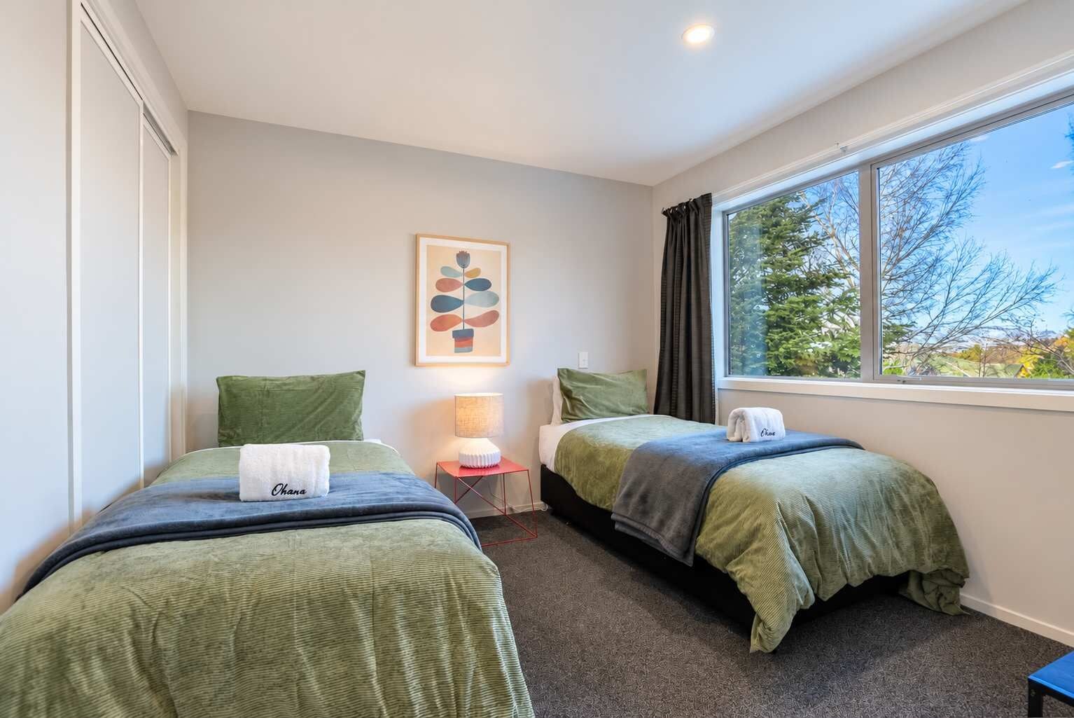 Een hotelkamer met twee bedden, een nachtkastje met een lamp, een kunstwerk in een lijst aan de muur, en een raam met uitzicht op bomen.
