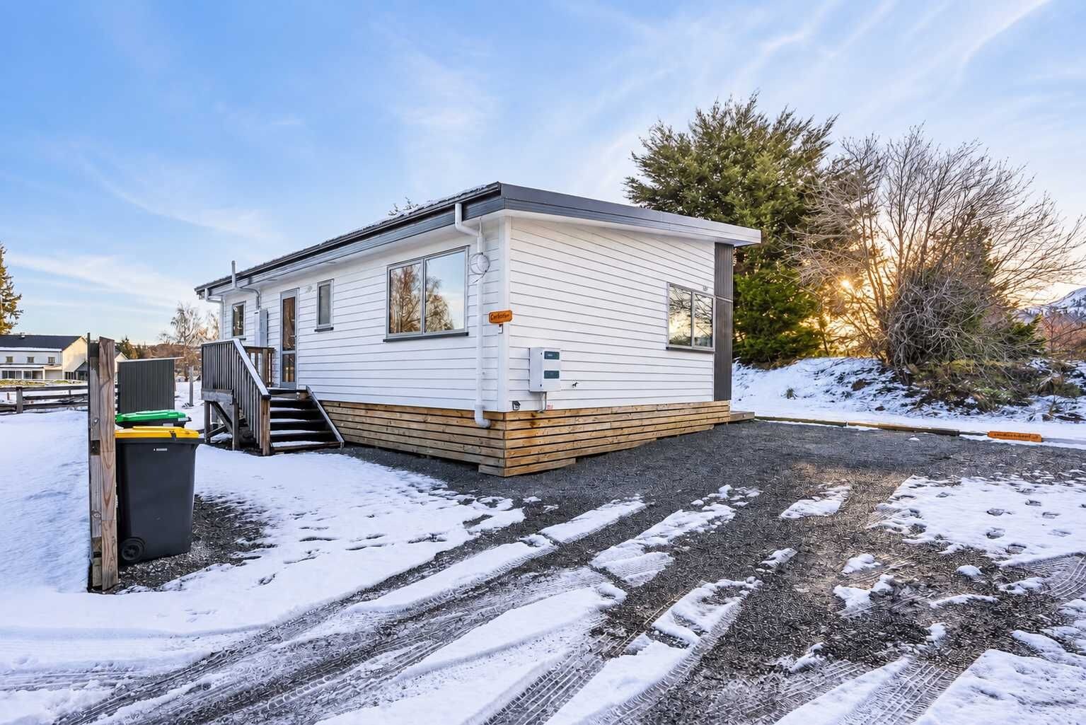 A single-story mobile home with a front porch, a small deck, and a driveway with visible tire tracks.