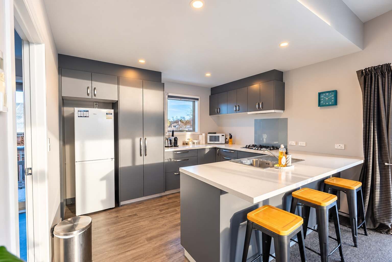 A modern kitchen with grey cabinets, a stainless steel refrigerator, a microwave, and a sink. There are three stools at the island.