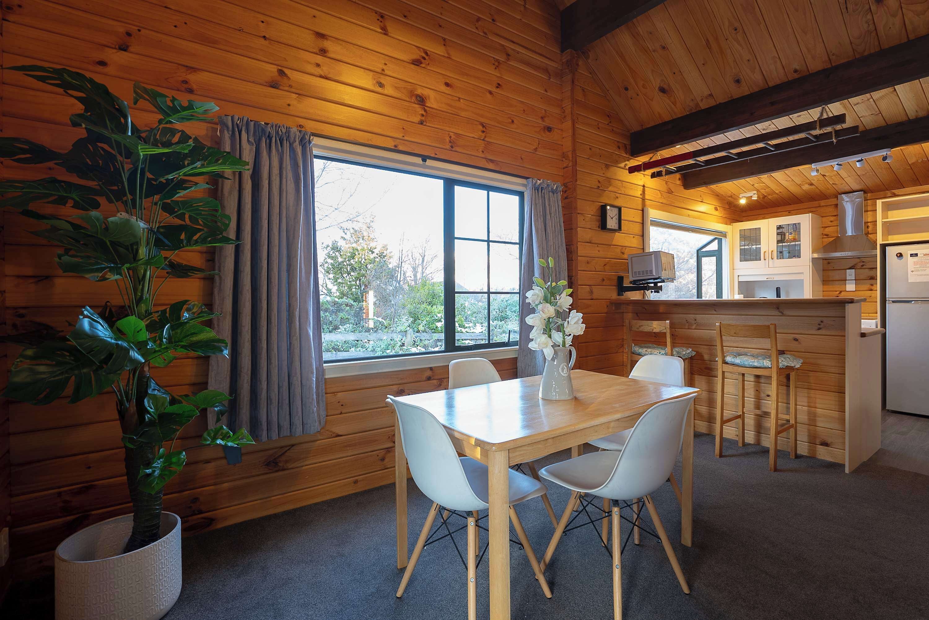 A wooden cabin interior with a dining table, chairs, a potted plant, a kitchen area, and a window with curtains.