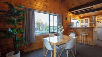 A wooden cabin interior with a dining table, chairs, a potted plant, a kitchen area, and a window with curtains.