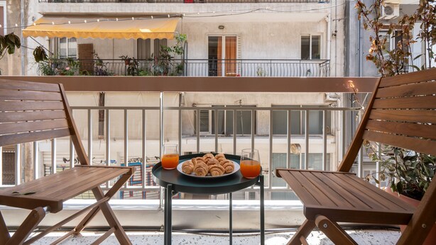 Un balcon avec deux chaises en bois, une petite table sur laquelle se trouvent une assiette de pâtisseries et des verres de jus d’orange, et un auvent jaune.