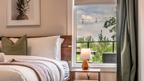 Quarto de hotel com cama, mesa de cabeceira com abajur, janela com vista para vegetação e uma ilustração de abacaxi emoldurada na parede.