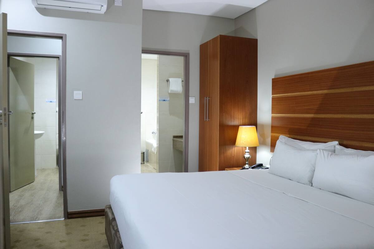 A hotel room with a bed, a wooden wardrobe, a bedside lamp, and a bathroom visible through an open door.