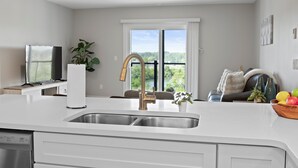 A modern kitchen with a stainless steel sink, a built-in dishwasher, and a large window offering a view of greenery. - Private Condo | CDGA Lake | Seasonal Hot Tub/Pool (Canandaigua)