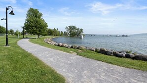 A paved path alongside a lake with boats in the distance, a lamp post, and a large tree. - Private Condo | CDGA Lake | Seasonal Hot Tub/Pool (Canandaigua)