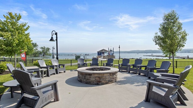 An outdoor seating area with Adirondack chairs and a stone fire pit, overlooking a body of water and a distant shoreline. - Private Condo | CDGA Lake | Seasonal Hot Tub/Pool (Canandaigua)