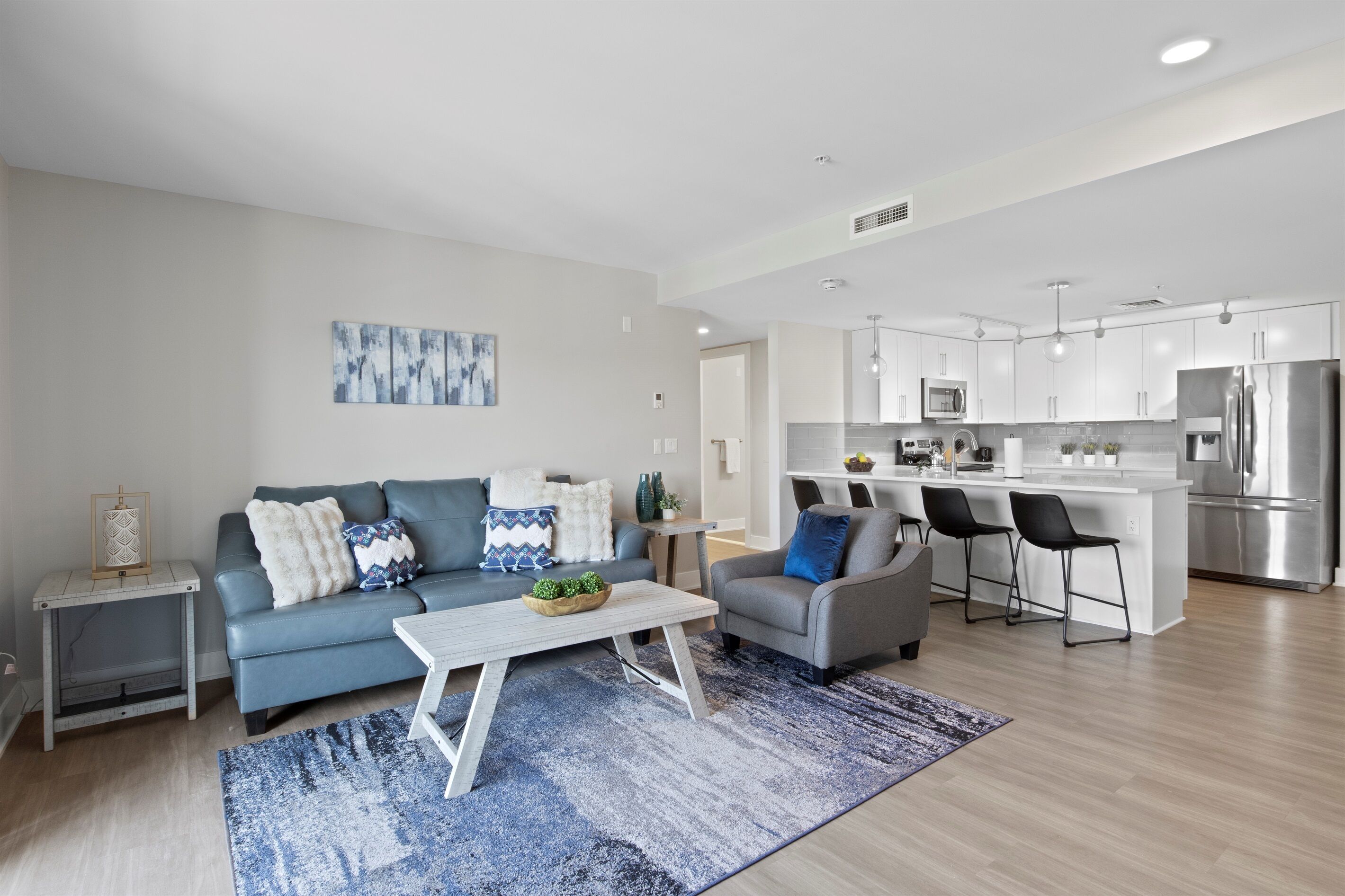 A modern living room with a sofa, coffee table, and a small side table. There is a kitchen area with a refrigerator, microwave, and bar stools.