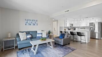 A modern living room with a sofa, coffee table, and a small side table. There is a kitchen area with a refrigerator, microwave, and bar stools.