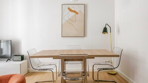 Un comedor moderno con una mesa de madera, sillas transparentes, un cuadro minimalista, una lámpara y un televisor sobre un mueble blanco.