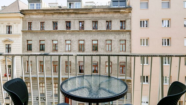 Un balcón con una mesa redonda y dos sillas, con vista a una hilera de edificios con ventanas y balcones uniformes.