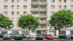 Un edificio residencial de varios pisos con balcones, coches aparcados y algunos árboles en la parte delantera.