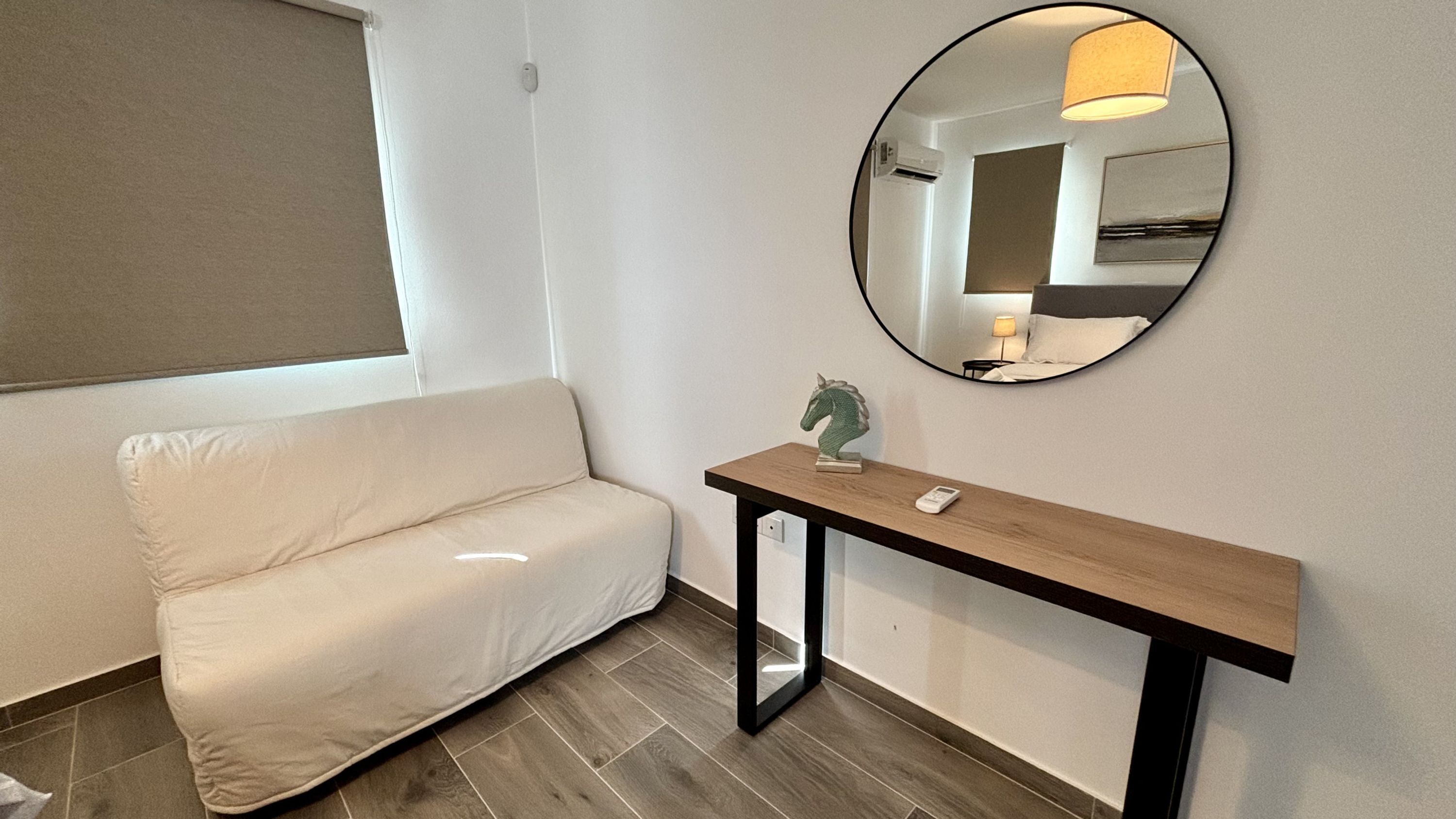 Quarto de hotel moderno com sofá, mesa consola com cabeça de cavalo decorativa e espelho redondo na parede.