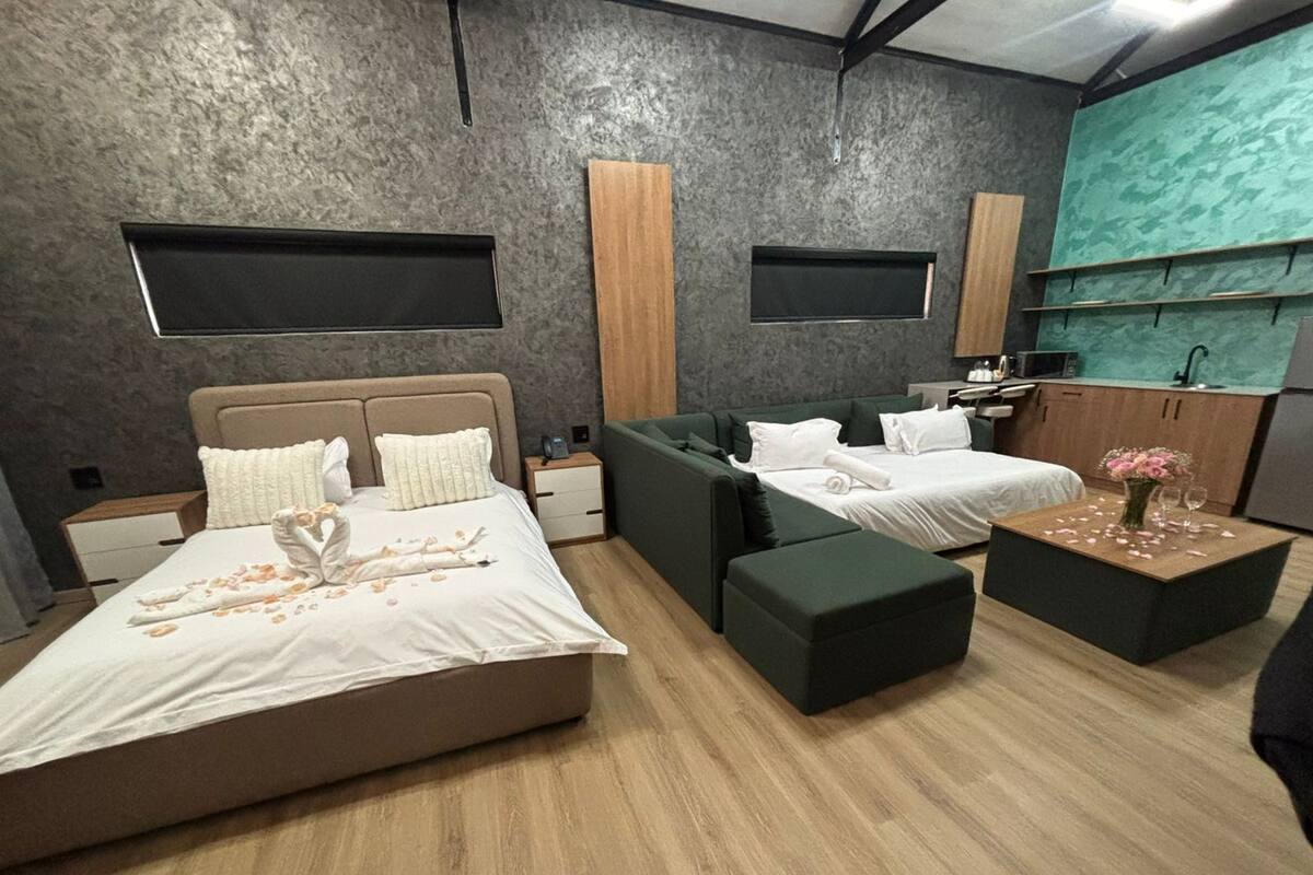 A modern hotel room with a large bed, a sofa, a small table, and a kitchenette.
