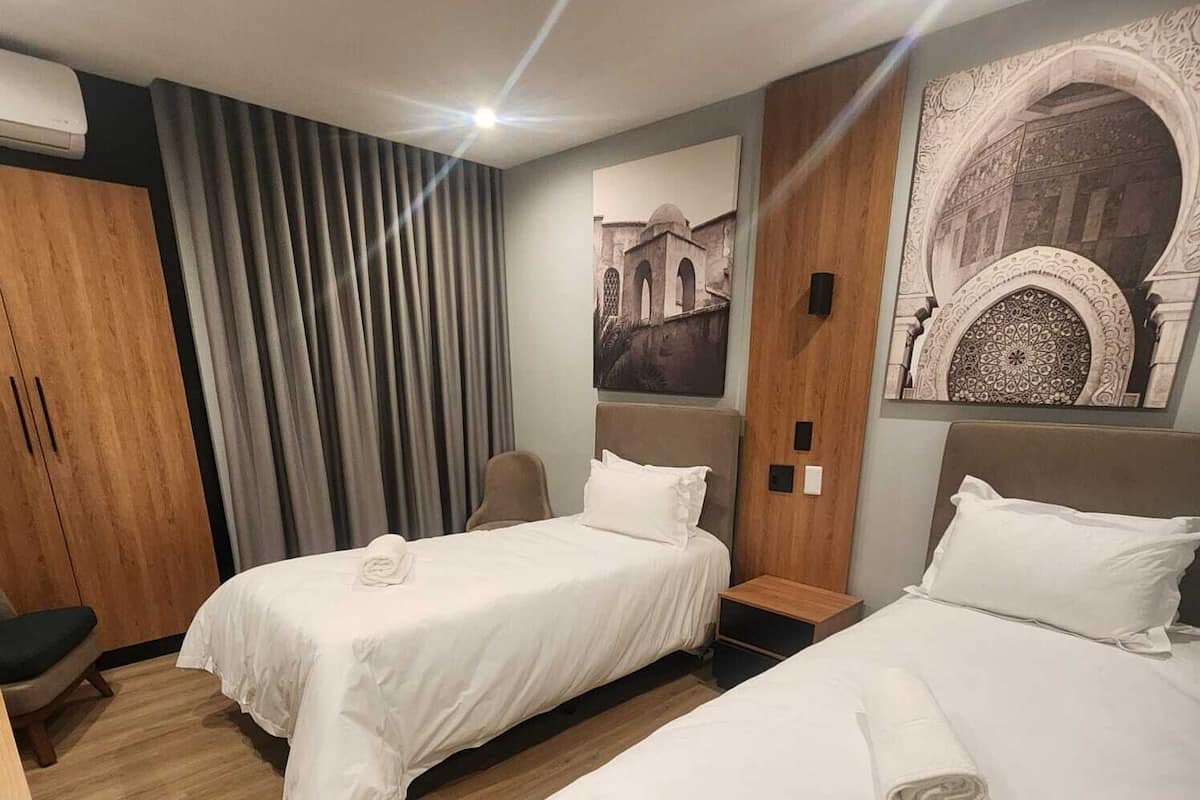 A hotel room with two beds, wooden flooring, a large window with curtains, a wall-mounted air conditioner, and a decorative wall piece.