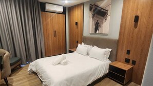 A hotel room with a bed, a chair, a wooden wardrobe, and a wall-mounted air conditioner. - The Lakewood Hotel  (Johannesburg South)