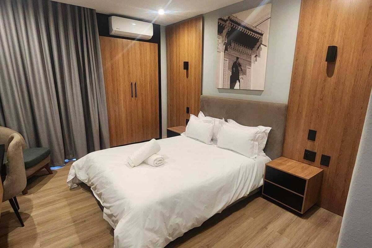 A hotel room with a bed, a chair, a wooden wardrobe, and a wall-mounted air conditioner.