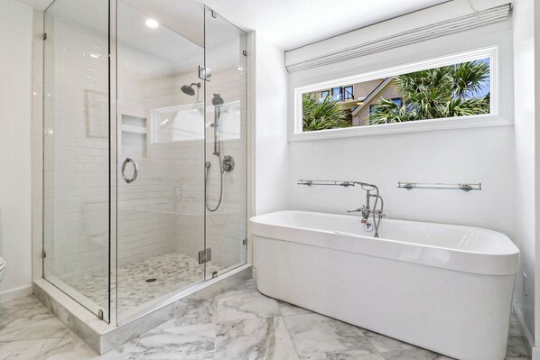 A modern bathroom with a glass-enclosed shower, a freestanding bathtub, and a window with a view of palm trees.