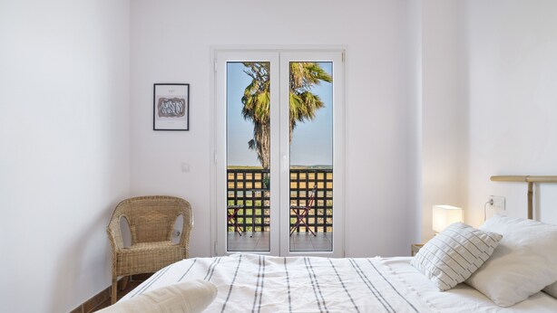 Ein Schlafzimmer mit einem Bett, einem Korbsessel, einer Tür mit Blick auf Palmen und einem gerahmten Bild an der Wand.