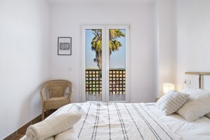 Un dormitorio con cama, una silla de mimbre, una puerta con vista a palmeras y un cuadro en la pared.