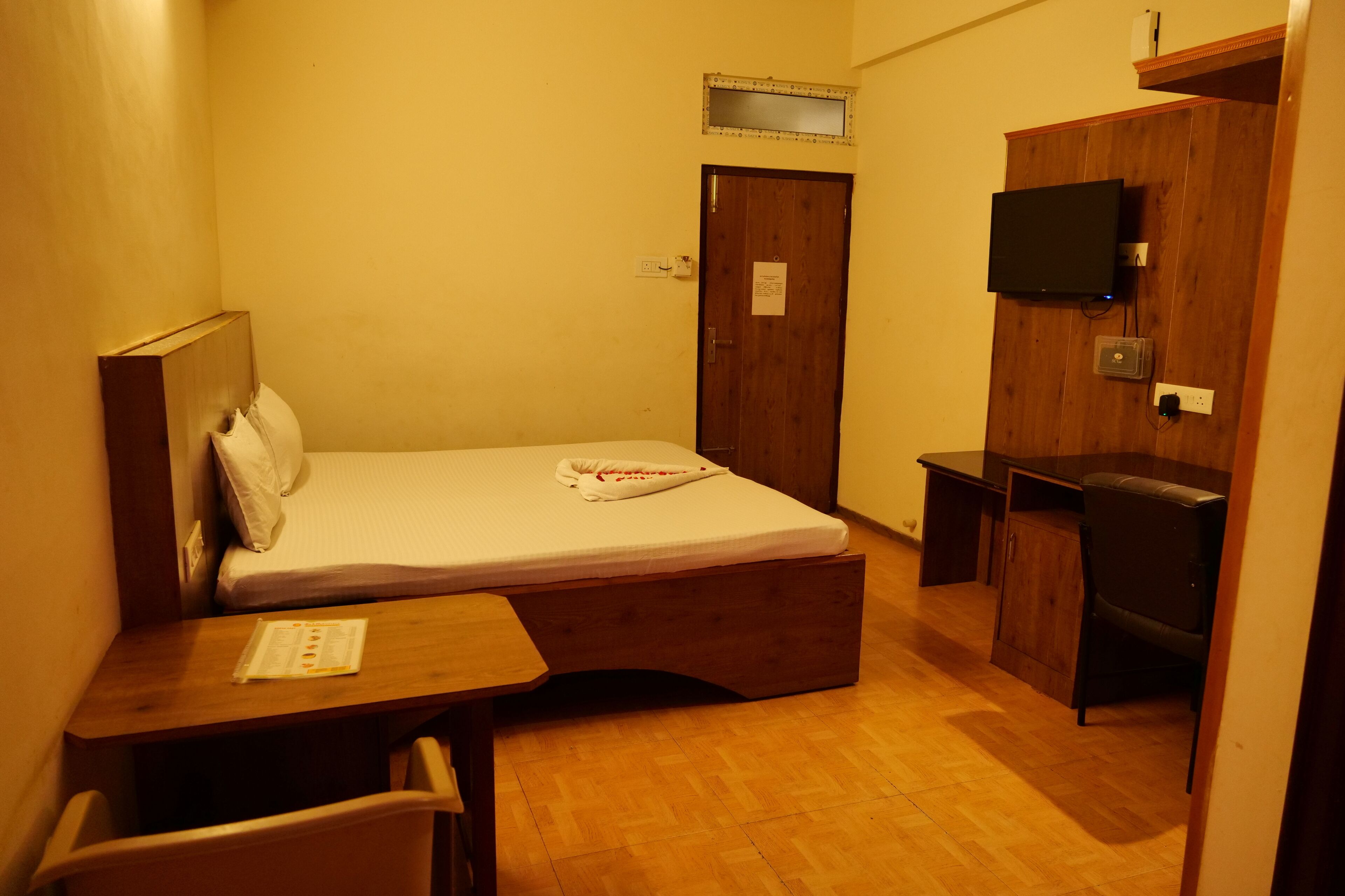 A compact hotel room with a single bed, a small desk, a chair, a TV mounted on the wall, and a wooden wardrobe.