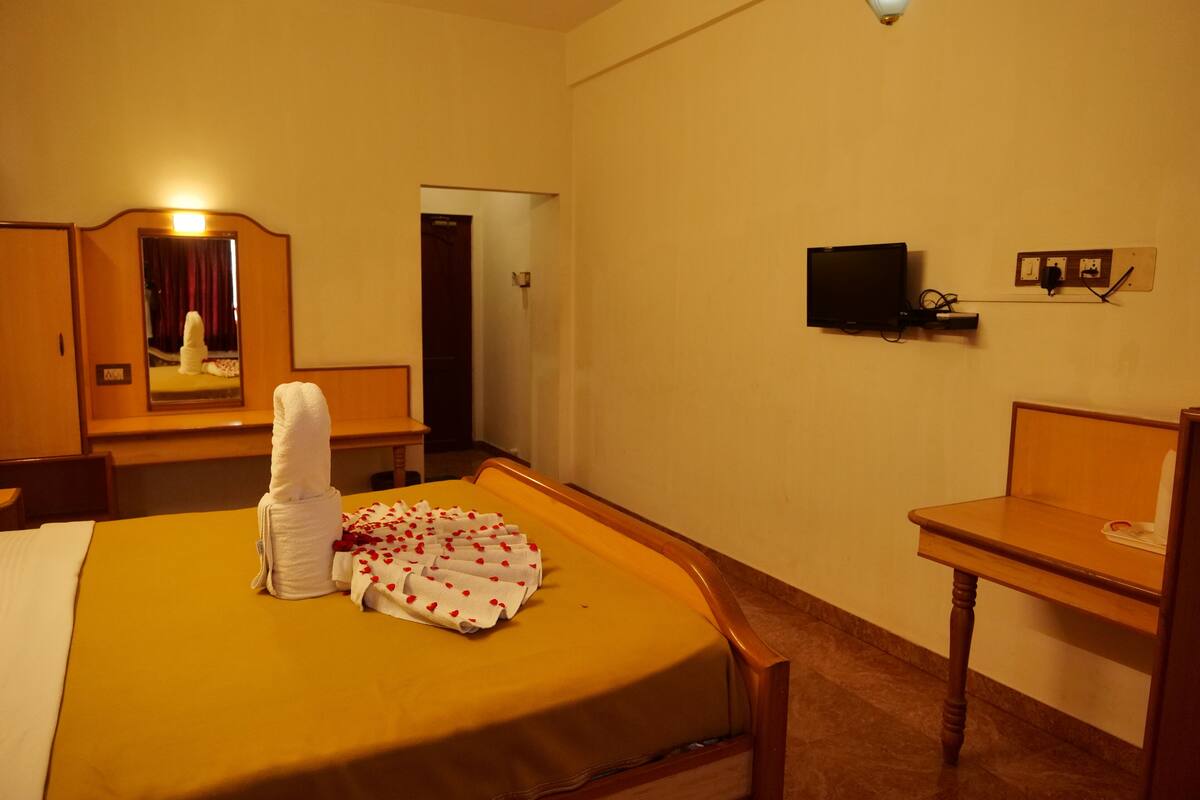A hotel room with a bed, a wooden nightstand, a television mounted on the wall, and a mirror above the bed.