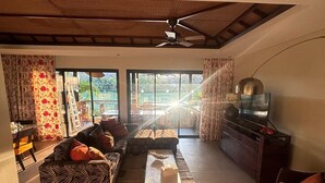 Un salón amplio con un sofá, una mesa de centro y una televisión. Cuenta con un ventilador de techo y vistas al exterior a través de amplias ventanas.