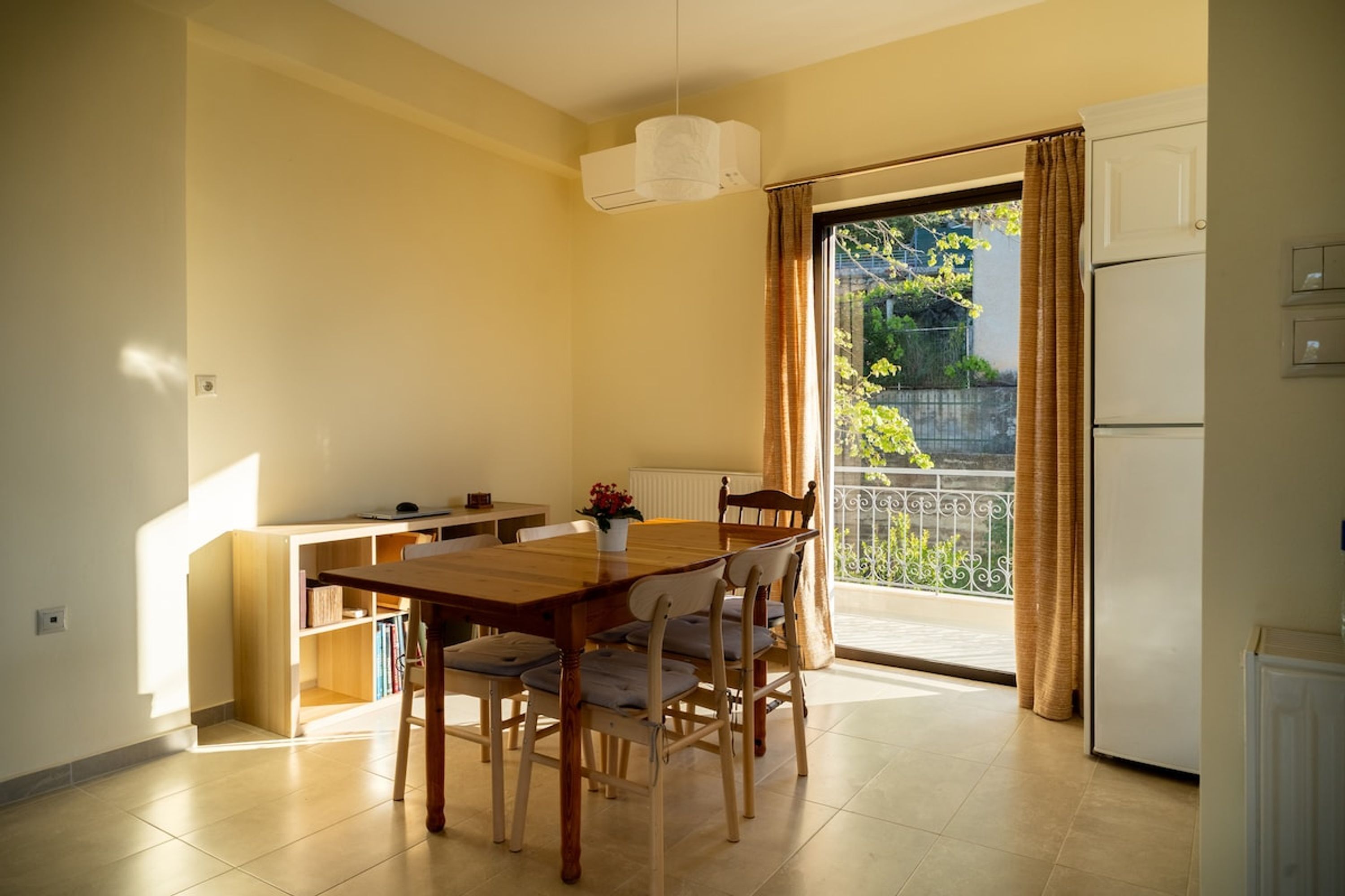A dining area with a wooden table and chairs, a small kitchen area with a refrigerator, and a view of a balcony with a railing and greenery.