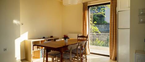 A dining area with a wooden table and chairs, a small kitchen area with a refrigerator, and a view of a balcony with a railing and greenery.
