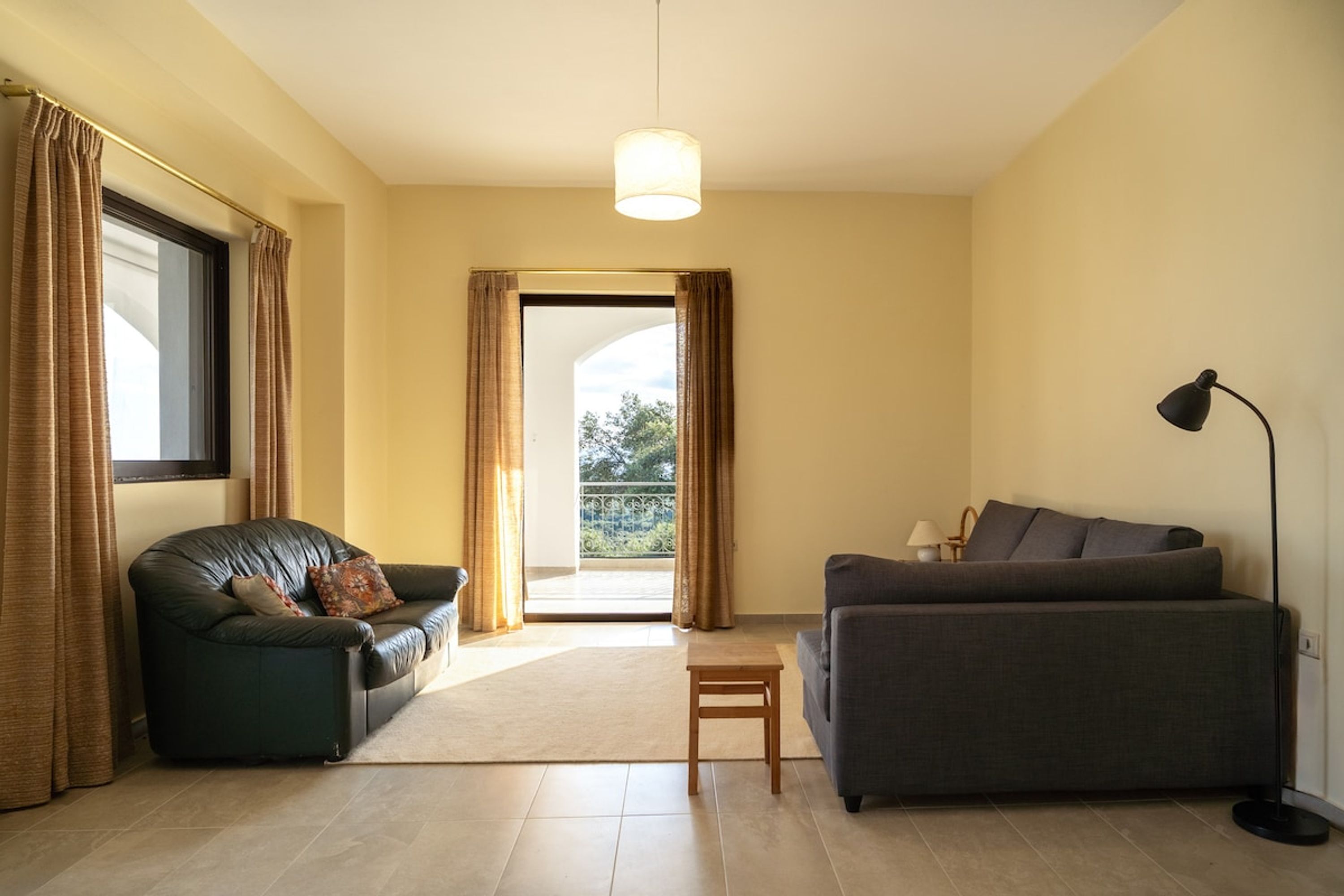 Una sala de estar con dos sofás, una mesita y una lámpara de pie. Se ve una ventana con cortinas, una puerta que da al exterior y vistas a la vegetación.