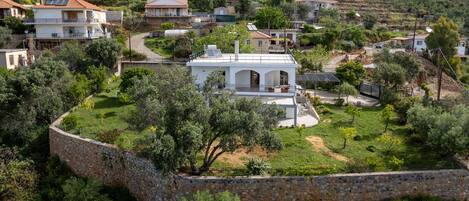 A white villa with a garden, surrounded by a stone wall, set in a hilly area with other houses.