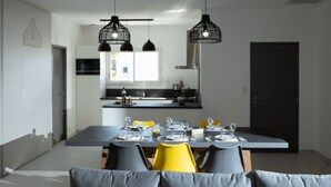 Un comedor moderno con una mesa grande dispuesta para seis personas, dos lámparas colgantes y una cocina al fondo.