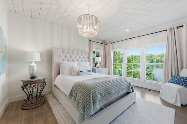 A bedroom with a large bed, a chandelier, a nightstand, a window with a view, and a chair.