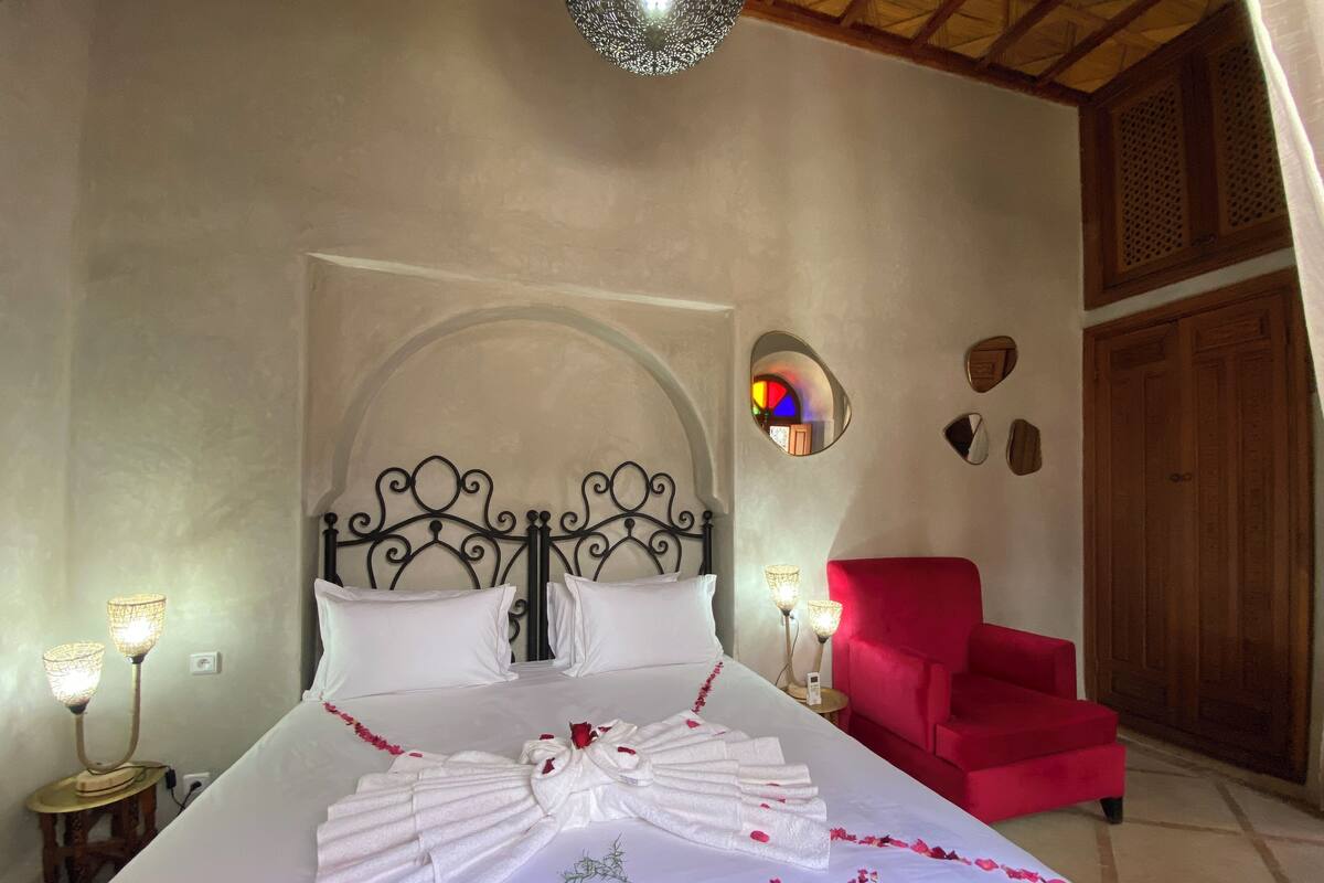 A neatly made bed with white linens and a decorative bow, a red armchair, and a small table with a lamp.