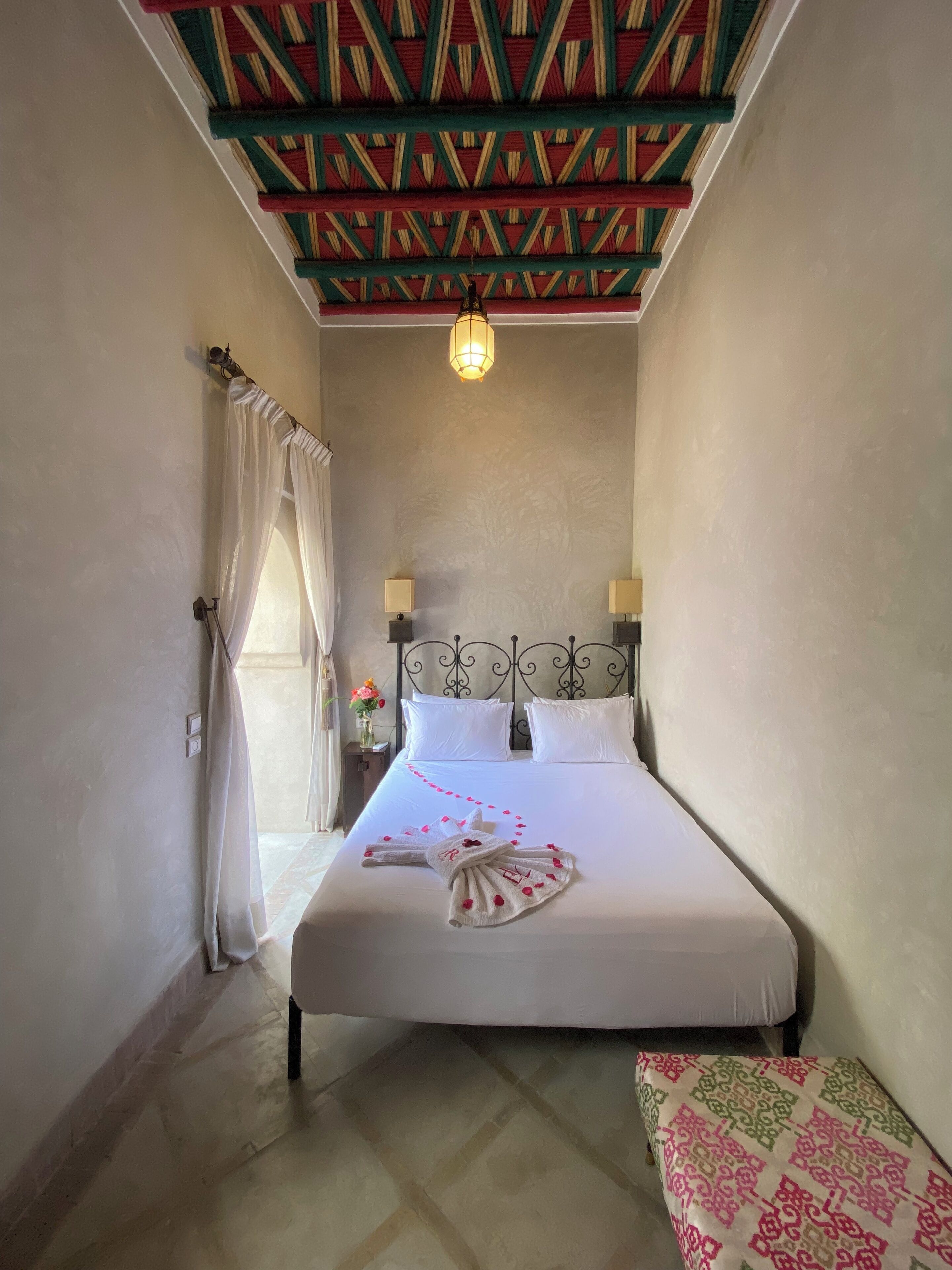 Un dormitorio con una cama, una mesita de noche con lámpara, un techo decorativo y un banco con cojín estampado.