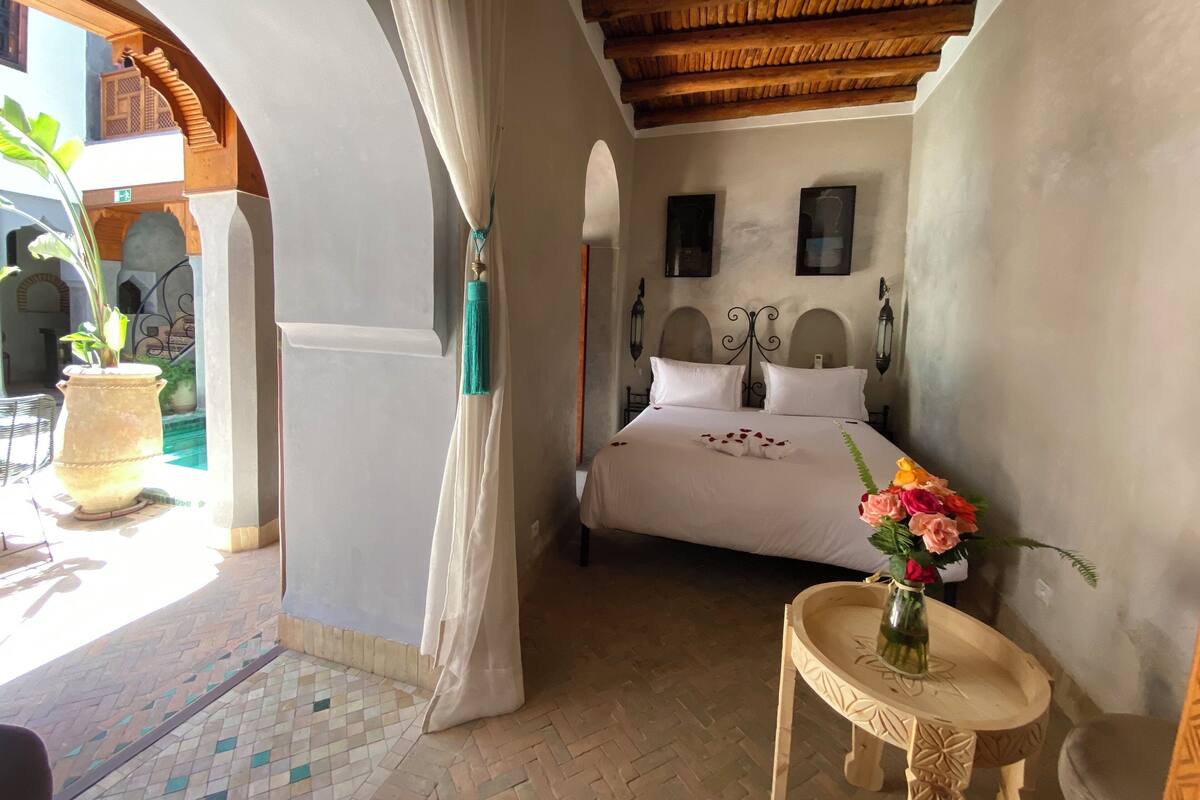 A bedroom with a bed, a small table with a vase, and a view of a courtyard.