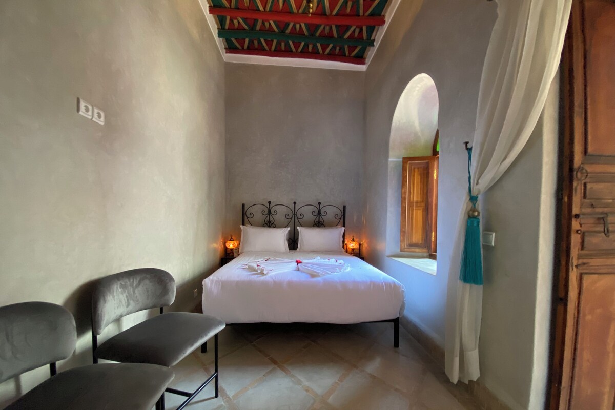 A bedroom with a bed, two chairs, a window, and a wooden door.