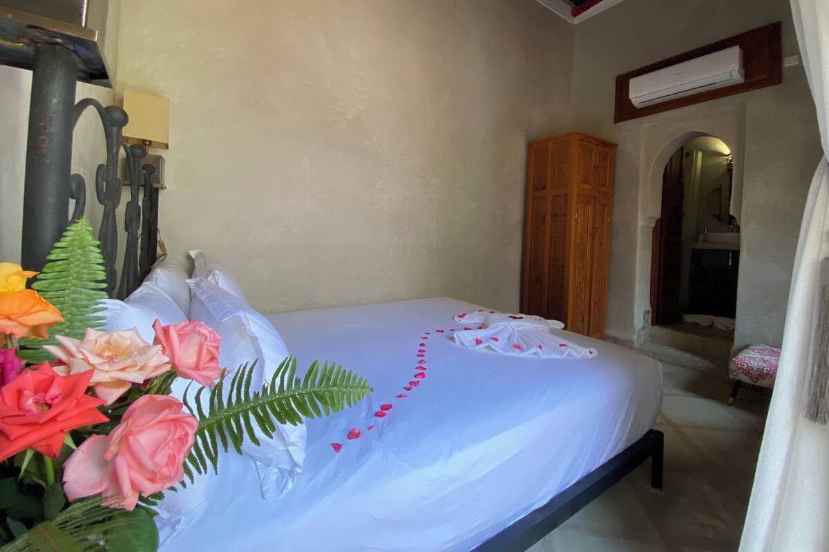 A neatly made bed with white linens, a wooden wardrobe, and a bathroom visible through an archway.