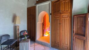 A room with a chair, a lamp, a small table with a vase, and a large wooden door with intricate carvings. - Riad Bab Yacout (Marrakech)