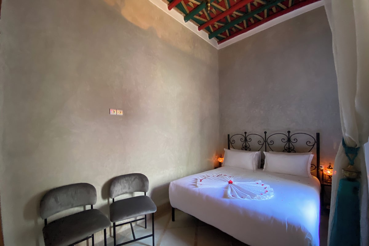 A bedroom with a bed, two chairs, a nightstand with a lamp, and a ceiling fan.