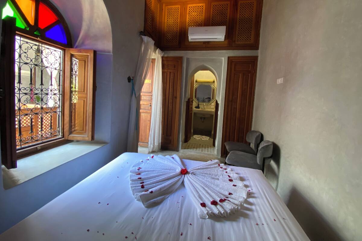 A room with a bed, a window with a stained glass design, a wooden ceiling, and a bathroom visible through an archway.