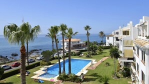 A pool area with palm trees, a clear view of the sea, and white buildings with balconies.