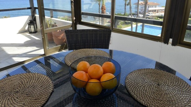 A glass table with a bowl of oranges, wicker chairs, and a view of the sea and palm trees.