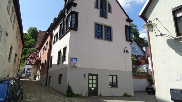 A narrow cobblestone street with buildings, a blue car parked on the left, and a building with a sign indicating a business. - Apartment 'Weitblick' (Top Floor Center) with Free Wi-Fi (max. 6 guests) (Pottenstein)