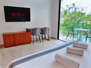 A hotel room with a bed, a TV mounted on the wall, a desk with two chairs, a small table, and a view of a pool and greenery outside.