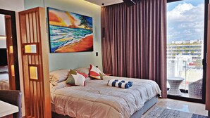 A hotel room with a bed, a painting of a beach, a view of the city, and a balcony with wicker furniture. - Centric Studio in 4th Floor with Balcony (Playa del Carmen)
