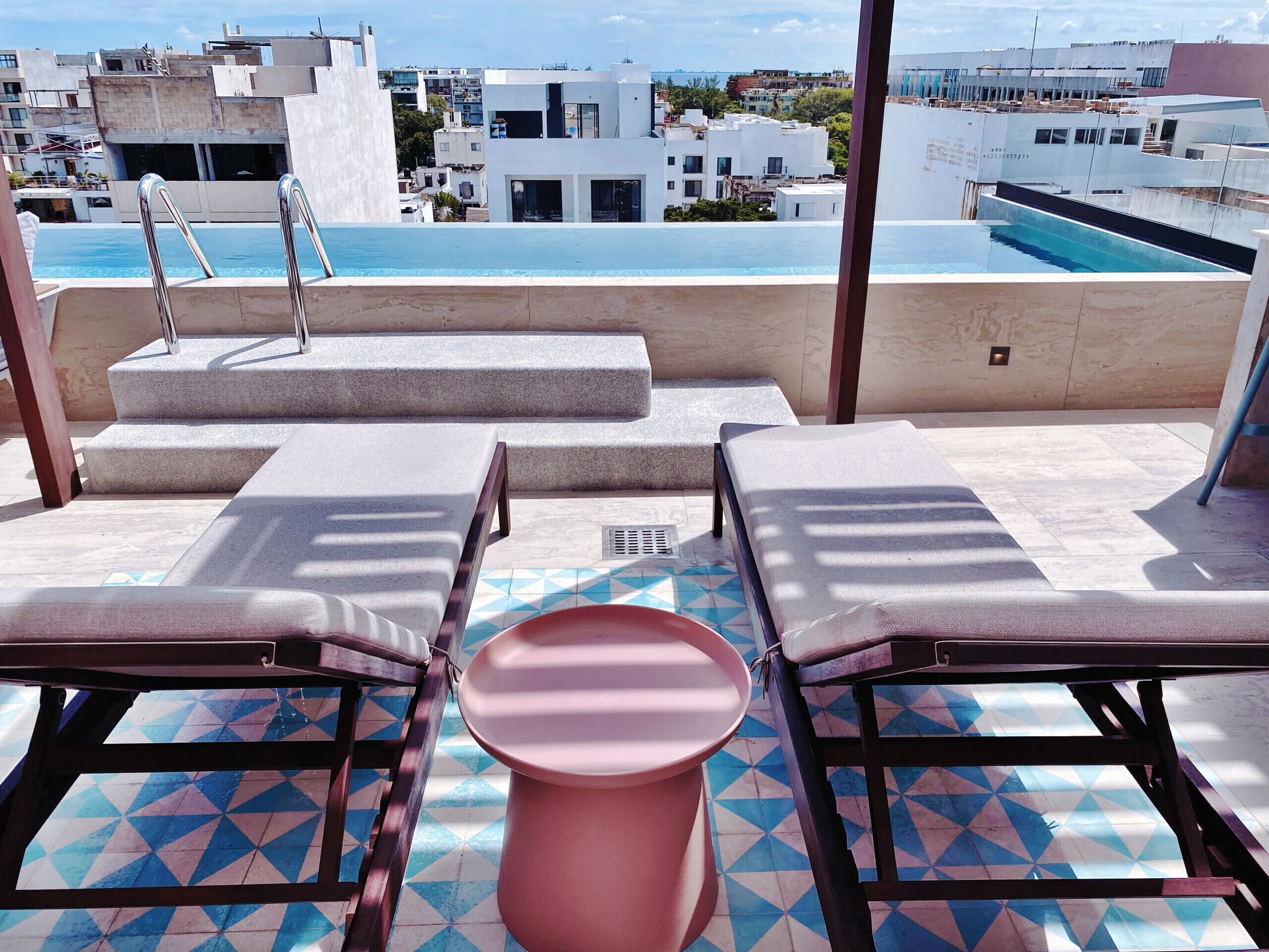 A rooftop pool area with two sun loungers, a pink table, and a view of the cityscape.