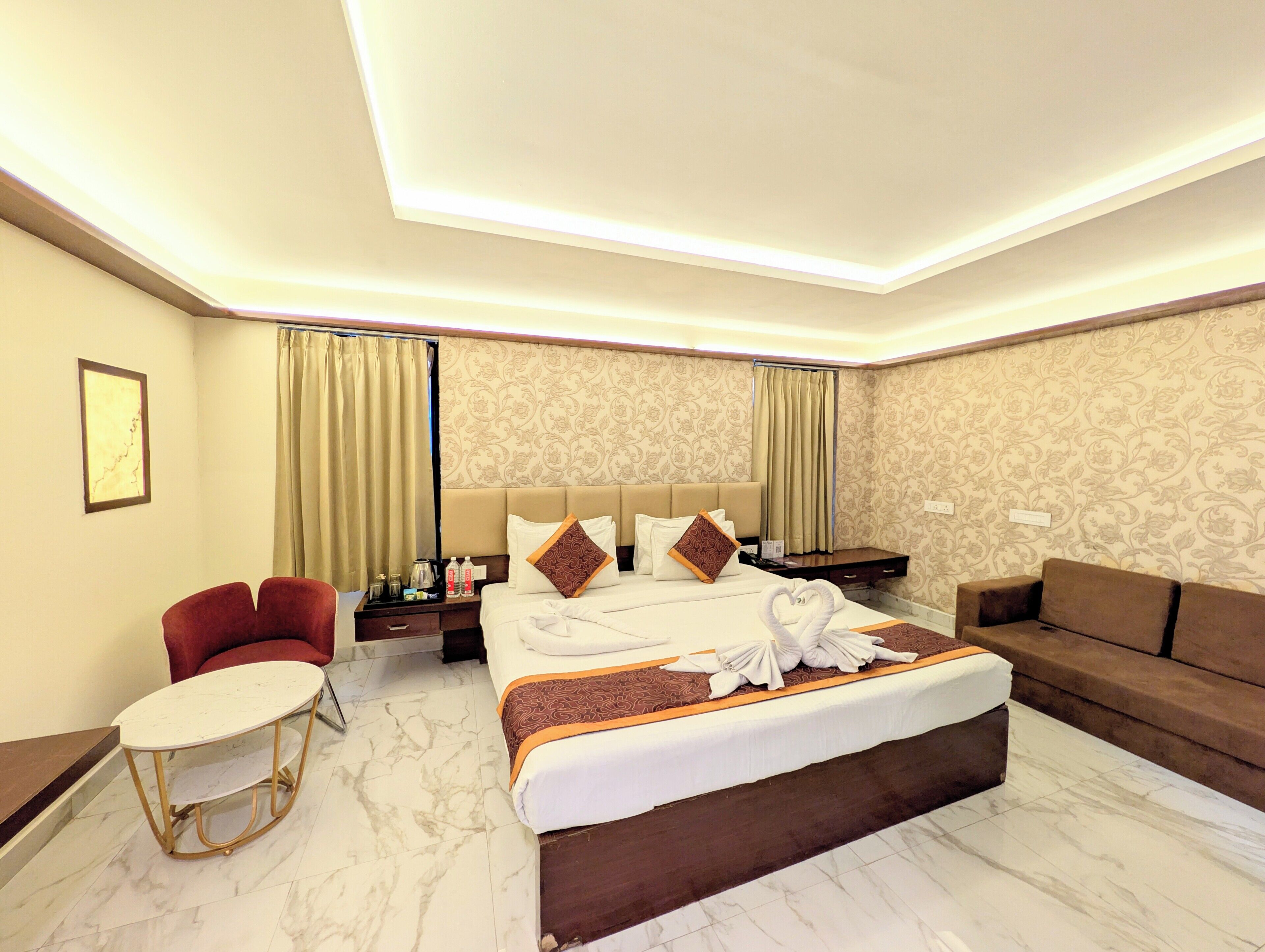 A hotel room with a bed, a red armchair, a small round table, a sofa, and a nightstand with a lamp.